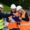 Auf dem Bild sind drei bautechnische AssistentInnen mit Schutzhelmen und Warnwesten zu sehen, die auf einer Baustelle Baupläne besprechen.