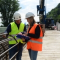 Auf dem Bild sind zwei bautechnische AssistentInnen zu sehen, die auf einer Baustelle Pläne besprechen, während sie Schutzhelme und Warnwesten tragen.