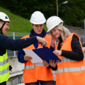 Auf dem Bild sind drei Personen zu sehen, die Schutzhelme und Warnwesten tragen und auf einer Baustelle Baupläne oder Dokumente besprechen.