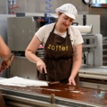 Zwei Chocolatiers/-ières arbeiten in einer Schokoladenmanufaktur und verteilen flüssige Schokolade auf einer Fläche.