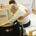 Eine Pflegeassistentin unterstützt eine ältere Person beim Transfer vom Bett in einen Rollstuhl.