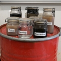 Das Bild zeigt mehrere Probengefäße mit unterschiedlichen Flüssigkeiten und Beschriftungen, die in der Entsorgungs- und Recyclingtechnik verwendet werden.