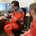 Auf dem Bild sind Entsorgungs- und RecyclingtechnikerInnen in orangefarbener Arbeitskleidung zu sehen, die in einem Büro mit Computern und Überwachungsbildschirmen arbeiten.