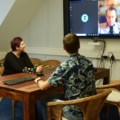 Zwei GamedesignerInnen sitzen in einem Besprechungsraum und führen eine Videokonferenz mit einer dritten Person auf einem Bildschirm.