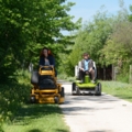 Zwei Garten- & GrünflächengestalterInnen fahren auf Aufsitzrasenmähern eine von Bäumen gesäumte Straße entlang.