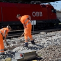 Zwei GleisbautechnikerInnen arbeiten in orangener Schutzkleidung an einem Bahngleis.