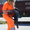 Eine Gleisbautechnikerin in Sicherheitskleidung arbeitet mit einem schweren Werkzeug auf einer Baustelle.