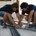 Das Bild zeigt zwei Personen, die an der Installation von Kabeln oder Rohren in einem Gebäude arbeiten.