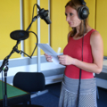 Eine Journalistin steht mit Kopfhörern vor einem Mikrofon in einem Tonstudio und liest von einem Blatt ab.