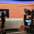 Das Bild zeigt ein Fernsehstudio mit einem Moderator, der an einem Schreibtisch steht, während Kameras auf ihn gerichtet sind.