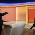 Das Bild zeigt einen Moderator, der in einem Fernsehstudio vor einer Kamera steht.