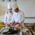 Das Bild zeigt zwei Köche in einer professionellen Küche, die gemeinsam an einem Gericht arbeiten.