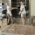 In dem Bild sind zwei Konditorinnen zu sehen, die in einer Bäckerei oder Konditorei an einem großen Ofen arbeiten und frische Backwaren zubereiten.