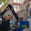 Ein Kran- und Baumaschinenführer bedient einen LKW-Kran auf einer Baustelle vor einem historischen Gebäude.