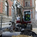Auf dem Bild ist ein Kran- und Baumaschinenführer zu sehen, der einen Bagger bedient, um Erdarbeiten vor einem historischen Gebäude durchzuführen.