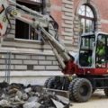 Auf dem Bild ist ein Baumaschinenführer zu sehen, der einen Bagger benutzt, um Schutt vor einem Gebäude zu bewegen.