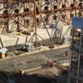 Das Bild zeigt eine Baustelle, auf der Bauarbeiter an einer Konstruktion aus Stahlträgern und Beton arbeiten. Das Bild ist wahrscheinlich von einem Kran aus aufgenommen und zeigt die Perspektive eines Kranführers/einer Kranführerin.