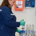 Eine Person führt im Labor mit Schutzkleidung und Handschuhen eine Pipettierarbeit durch, was typisch für den Beruf einer Labortechnikerin in der Biochemie oder Chemie ist.