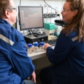 Das Bild zeigt zwei Labortechnikerinnen in Schutzkleidung, die vor einem Computer in einem Labor sitzen und sich mit chemischen Proben beschäftigen.