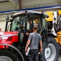 Zwei Land- und Baumaschinentechniker arbeiten in einer Werkstatt an landwirtschaftlichen Maschinen, darunter ein Traktor.