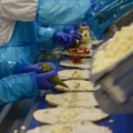 Das Bild zeigt LebensmitteltechnikerInnen in Schutzkleidung, die an einem Fließband Pizzen mit verschiedenen Zutaten belegen.