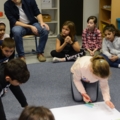 Ein Lehrer sitzt und beaufsichtigt eine Gruppe von Kindern, die gemeinsam auf dem Boden an einem Projekt arbeiten.