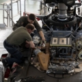 Das Bild zeigt zwei Luftfahrzeugtechniker, die mit der Wartung oder Reparatur des Rotorsystems eines Hubschraubers beschäftigt sind.