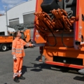 Auf dem Bild ist eine Müllauflegerin in orangefarbener Arbeitskleidung zu sehen, die an einem Müllwagen arbeitet.