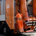 Auf dem Bild ist eine Müllarbeiterin zu sehen, die in orangefarbener Schutzkleidung auf einem Müllwagen steht.