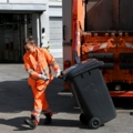 Das Bild zeigt eine Müllauflegerin in orangener Arbeitskleidung, die einen Müllcontainer zu einem Müllwagen schiebt.