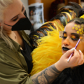 Das Bild zeigt eine Maskenbildnerin, die detailreiches Make-up auf das Gesicht einer Person aufträgt, die ein auffälliges Kostüm trägt.