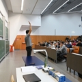 Ein Dozent für Mathemarik steht vor einer gefüllten Universitätsvorlesung, während er an einer Tafel erklärt.
