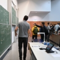 Auf dem Bild ist ein Hörsaal zu sehen, in dem eine Vorlesung gerade ihr Ende findet und ein Student den Hochschullehrer noch eine Frage zu einer mathematischen Formel stellt.