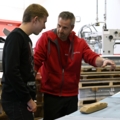 Ein Medienfachmann erklärt einem Kollegen in einer Werkstatt etwas über ein Holzobjekt.