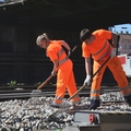 Auf dem Bild sind zwei Personen in orangener Arbeitskleidung zu sehen, die auf Bahngleisen mit Werkzeugen arbeiten.
