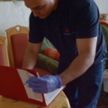 Ein Pflegeassistent in Arbeitskleidung überprüft Unterlagen in einem Ordner, während eine ältere Person im Hintergrund sitzt.