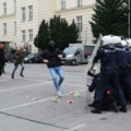 Auf dem Bild sind Exekutivbedienstete im Polizeidienst in Schutzausrüstung zu sehen, die sich gegen eine Gruppe von Demonstrierenden verteidigen.