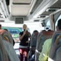 Auf dem Bild spricht eine Reiseleiterin mit einem Mikrofon zu den Passagieren in einem Reisebus.