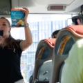 Auf dem Bild hält eine Reiseleiterin in einem Reisebus ein Mikrofon und eine Tüte "Wiener Zucker" hoch, während sie eine Gruppe von Reisenden informiert.