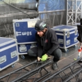 Ein Veranstaltungstechniker mit Helm und Handschuhen montiert Bühnentechnik auf einem Konzert.