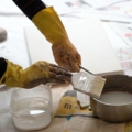 Eine Person trägt gelbe Handschuhe und arbeitet mit Pinsel und Farbe.