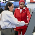 Eine Flughafenmitarbeiterin in roter Uniform hilft einem Passagier am Check-in-Automaten.