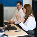 Das Bild zeigt zwei Personen in einem Büro, die an Computern arbeiten und sich möglicherweise in einer Besprechung befinden.
