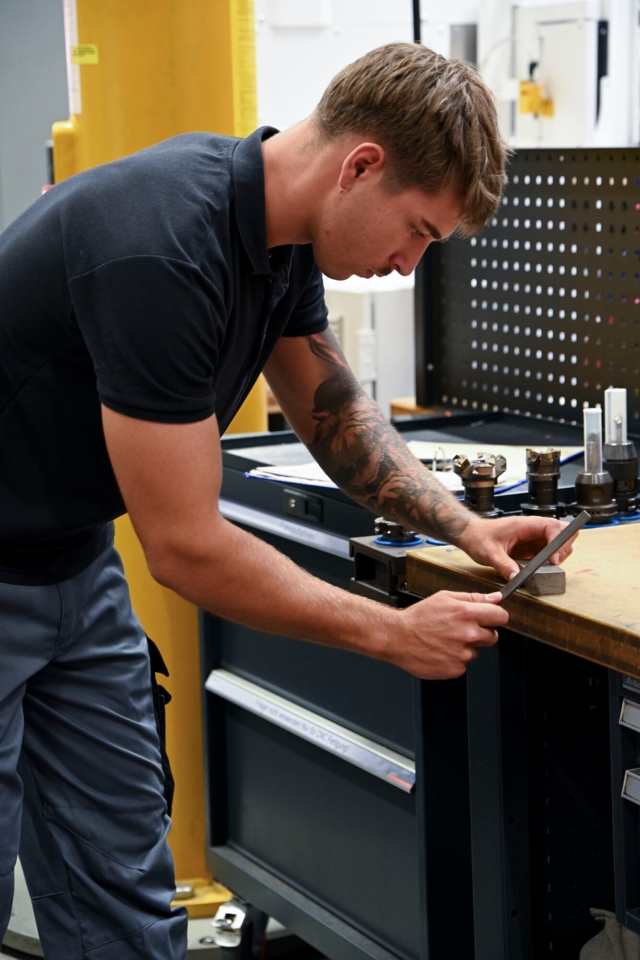 Ein Mann arbeitet mit einer Feile an einem Metallstück in einer Werkstatt.