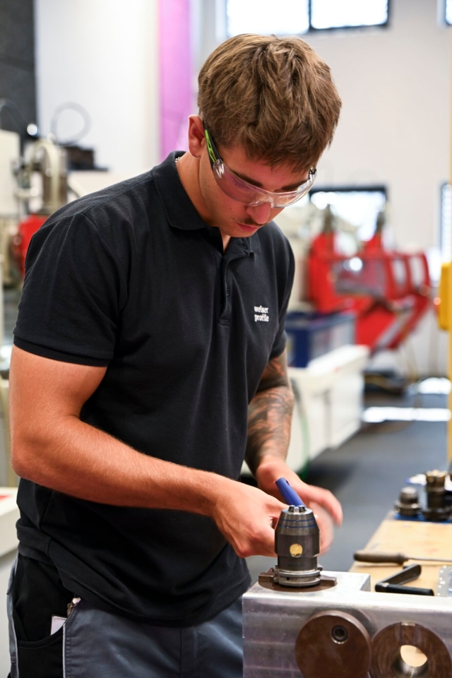Auf dem Bild ist eine Person zu sehen, die in einer Werkstatt mit einem Werkzeug an einem Metallstück arbeitet.
