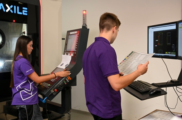 Zwei Personen arbeiten an einer computergesteuerten Maschine und überprüfen technische Zeichnungen oder Pläne.