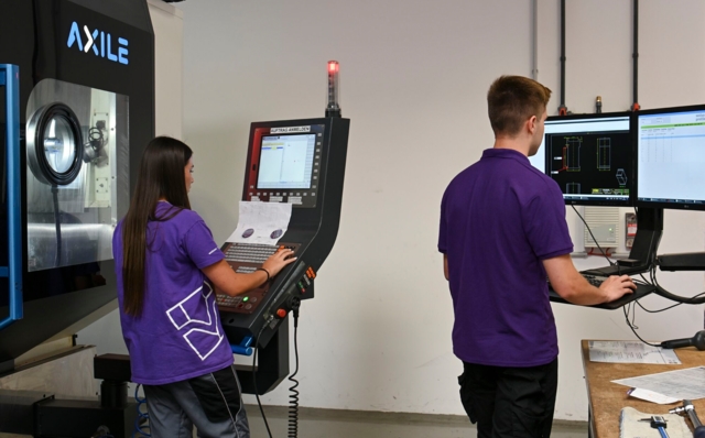 Zwei Personen bedienen Maschinen und Computer in einem industriellen Umfeld.