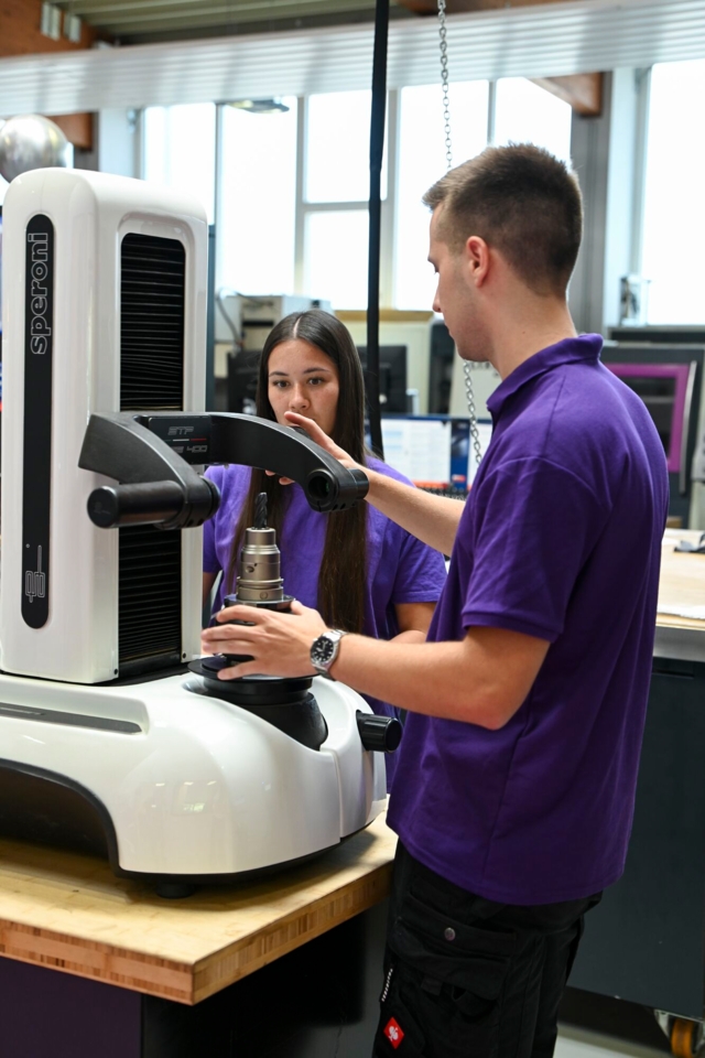 Zwei Personen bedienen in einem Werkstattumfeld eine industrielle Messmaschine oder Kalibrierungsgerät.